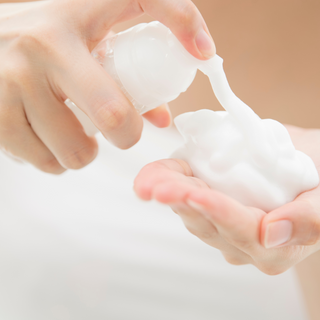 image is of foaming facial cleanser being used. The foam is coming out of a white bottle and is being foamed on the hands.