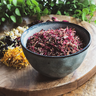 a pretty ceramic bowl sits on a wooden table. The bowl is filled with dried cornflower petals that are purple