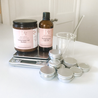 Image is of a lip balm kit with scales. There is a glass jar with butter, a glass amber bottle with oil, and lip balm pots and tins are in the front. There is a glass beaker and glass rod on the right.