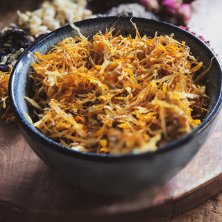 A bowl of bright yellow dried Calendula or Marigold sits on a timber table with other coloured dried flowers in the background