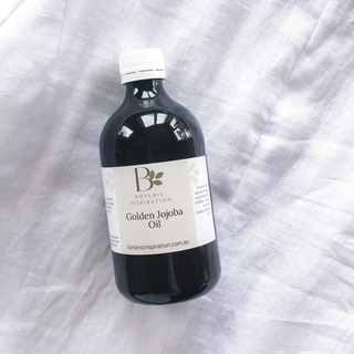 a bottle of golden jojoba oil lays on a white linen surface. the bottle is amber and has a cream and white label with the botanic inspiration logo