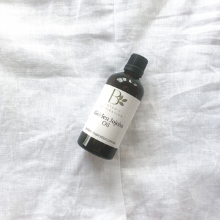 An amber bottle lays on a white linen surface. the label reads golden jojoba oil
