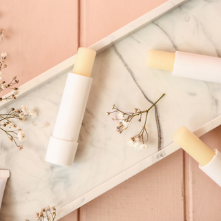 a close up of 3 vanilla flavoured lip balms in white tubes are on a white ceramic dish with a pink background. There are small white flowers decorating