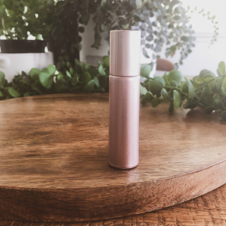 A pink opaque roller bottle sits on a timber surface with green plants in the background